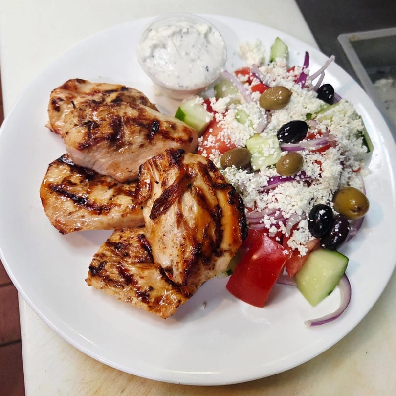 Grilled chicken pieces with a Mediterranean salad topped with feta, olives, and a side of tzatziki.