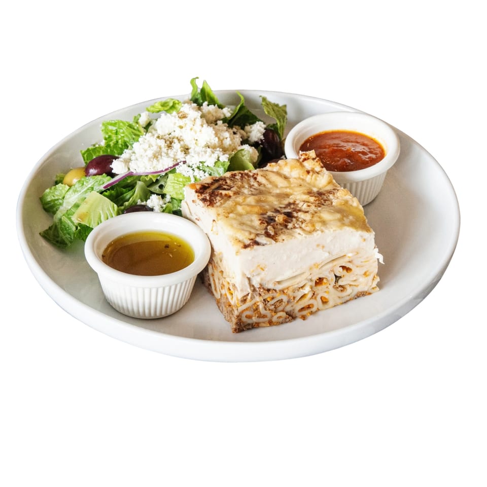 Pastitsio with a side salad topped with feta, olives, and two small bowls of dressing.